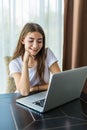 Young smiling girl is sitting on modern chair near the window in light cozy room at home working on laptop in relaxing atmosphere Royalty Free Stock Photo