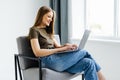 Young smiling woman sitting on modern chair near the window in light cozy room at home working on laptop in relaxing atmosphere Royalty Free Stock Photo