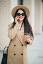 Young smiling business woman in sunglasses and coat stands near office centre with cup of coffee and talks on smart phone. Royalty Free Stock Photo