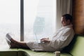 Young smiling businessman working on laptop computer wearing white bath robe sitting near window with cup of coffee Royalty Free Stock Photo