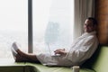 Young smiling businessman working on laptop computer wearing white bath robe sitting near window with cup of coffee Royalty Free Stock Photo