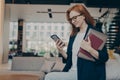 Young smiling businesswoman in glasses with laptop and agenda under her arm looking at smartphone Royalty Free Stock Photo