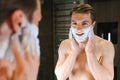 Young smiling Caucasian shirtless man applying shaving foam on face in front of mirror, preparing for hair removal Royalty Free Stock Photo