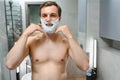 Young smiling Caucasian shirtless man applying shaving foam on face in front of mirror, preparing for hair removal Royalty Free Stock Photo