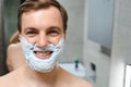Young smiling Caucasian shirtless man applying shaving foam on face in front of mirror, preparing for hair removal Royalty Free Stock Photo