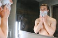 Young smiling Caucasian shirtless man applying shaving foam on face in front of mirror, preparing for hair removal Royalty Free Stock Photo