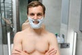 Young smiling Caucasian shirtless man applying shaving foam on face in front of mirror, preparing for hair removal Royalty Free Stock Photo