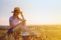 Young smiling caucasian woman in a hat uses a mobile phone sitting on the grass in a park at sunset. Happy girl writes a message Royalty Free Stock Photo