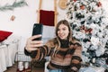 Young smiling caucasian woman making selfie using mobile phone near Christmas tree at home Royalty Free Stock Photo