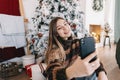 Young smiling caucasian woman making selfie using mobile phone near Christmas tree at home Royalty Free Stock Photo