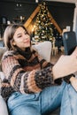 Young smiling caucasian woman using mobile phone, making selfie near Christmas tree at home Royalty Free Stock Photo