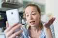 Young smiling cheerful pleased woman indoors at home kitchen using social media apps on mobile phone for chatting and Royalty Free Stock Photo