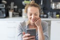 Young smiling cheerful pleased woman indoors at home kitchen using social media apps on mobile phone for chatting and Royalty Free Stock Photo