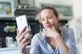 Young smiling cheerful pleased woman indoors at home kitchen using social media apps on mobile phone for chatting and Royalty Free Stock Photo