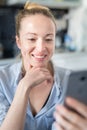 Young smiling cheerful pleased woman indoors at home kitchen using social media apps on mobile phone for chatting and Royalty Free Stock Photo