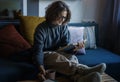 Young smiling cheerful woman in a warm sweater and eyeglasses  reading a book while sitting on the sofa with a mug of coffee Royalty Free Stock Photo