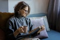 Young smiling cheerful woman in a warm sweater and eyeglasses  reading a book while sitting on the sofa with a mug of coffee Royalty Free Stock Photo