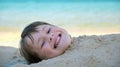 Young smiling child girl lying down covered with white sand on tropic beach on blue sky and ocean water background Royalty Free Stock Photo