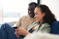 Young Smiling Couple Relaxing At Home Sitting On Sofa Checking Social Media On Mobile Phones Together Royalty Free Stock Photo