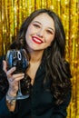 Young smiling cute woman with make-up wearing black shirt, holding glass with red wine on golden tinsel background. Royalty Free Stock Photo