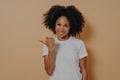 Young smiling dark skinned female curly hair pointing aside with thumb, isolated over beige wall
