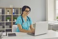 Young smiling doctor sitting and working on laptop online in medical clinic Royalty Free Stock Photo