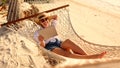 Young smiling female, happy woman in straw hat and sunglasses using digital tablet while relaxing in the hammock on tropical sandy Royalty Free Stock Photo