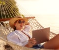 Young smiling female, happy woman in straw hat and sunglasses using digital tablet while relaxing in the hammock on tropical sandy Royalty Free Stock Photo