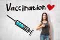 Young smiling girl giving thumbs up near wall with image of syringe and title `Vaccination` with cute little red heart. Royalty Free Stock Photo