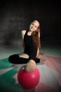 Young smiling girl gymnast in black sport body and uppers sitting on floor and touching pink gymnastic ball with leg