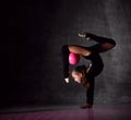 Young girl gymnast in black sport body and uppers standing in bridge pose and holding pink gymnastic ball between leg and neck