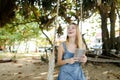 Young smiling girl using tablet and riding swing on sand, wearing jeans sundress. Royalty Free Stock Photo