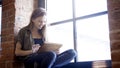 Young smiling happy attractive woman holding a digital tablet sitting on the window sill Royalty Free Stock Photo