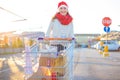 Young smiling happy woman with a lot of gift boxes in shopping cart going to her car in the mall parking wearing red Santa hat Royalty Free Stock Photo