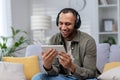 Young smiling hispanic man sitting on sofa at home wearing headphones and using tablet Royalty Free Stock Photo