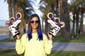 Young smiling informal girl with blue hair and retro sunglasses holding roller skates in her hands. Woman in the park with rollers