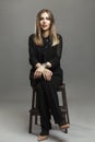 A young smiling lady in strict black clothes sits on a chair. Beautiful blonde in a black suit. Gray background. Vertical. Full Royalty Free Stock Photo