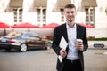 Young smiling man in black suit and white shirt with wireless earphones holding laptop and cup of coffe in hands while