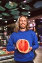 Young smiling man in dark blue sweater joyfully looking in camera while holding half of watermelon in hands in modern