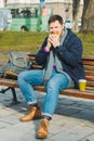 young smiling man eating burger and drink coffee sitting on city bench