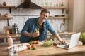 Young smiling man with glass of fresh juice cooking breakfast in the kitchen checking recipe in his laptop , sunday morning Royalty Free Stock Photo