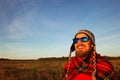 Young smiling man in the knitted cap, sunglasses and blanket is meeting of sunrise on the background of a field and blue sky. Royalty Free Stock Photo