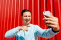 Young smiling man makes self portrait on smart phone on the background of red wall Royalty Free Stock Photo