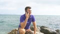 Young smiling man is talking on mobile phone on the beach in the sea. Handsome happy guy sitting on stone near the ocean Royalty Free Stock Photo