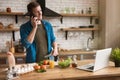 Young smiling man talking on the phone while cooking breakfast in the kitchen checking recipe in his laptop , sunday morning Royalty Free Stock Photo