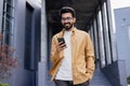 A young smiling man walks through the city with a phone in his hands, outside an office building, happily uses an