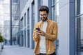 A young smiling man walks through the city with a phone in his hands, outside an office building, happily uses an