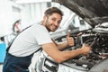 Young smiling mechanic in uniform fixing motor problems in car bonnet working in service center showing like sign Royalty Free Stock Photo