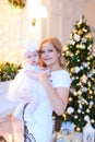 Young smiling mother wearing white dress and keeping little female baby near Christmas tree.