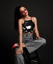 Young smiling pretty woman model in glittering top, jeans, red shoes and sunglasses sitting on stool and looking at Royalty Free Stock Photo